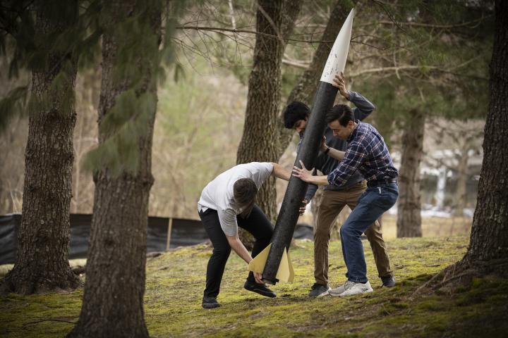 Three students hold up a large, black and white rocket, outside on Olin's campus. 