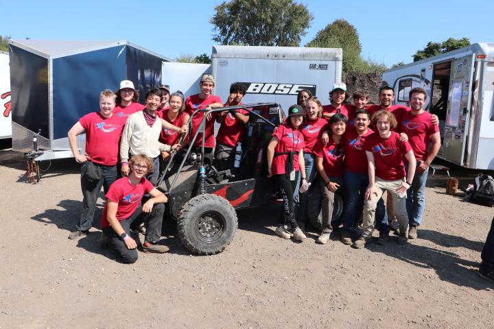 A large group of college students wearing red "Baja" t-shirts pose for a photo in front of a trailer.