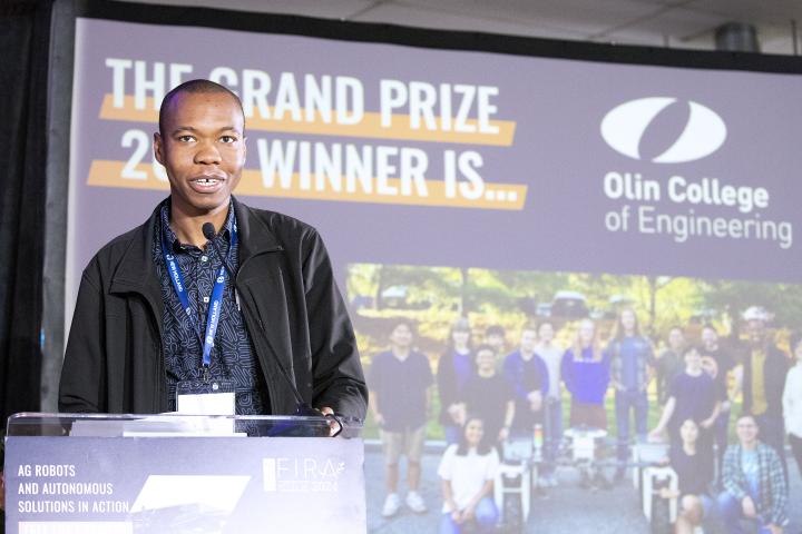 Olin College of Engineering's Kenechukwu Mbanisi, Assistant Professor of Robotics Engineering, is shown at a podium after the Olin teams wins the Farm Robotics Challenge. 