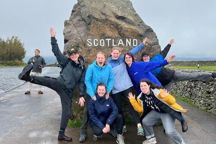 A group of college students pose for a joyful photo in front of a large rock with the words "SCOTLAND' across it in white lettering.