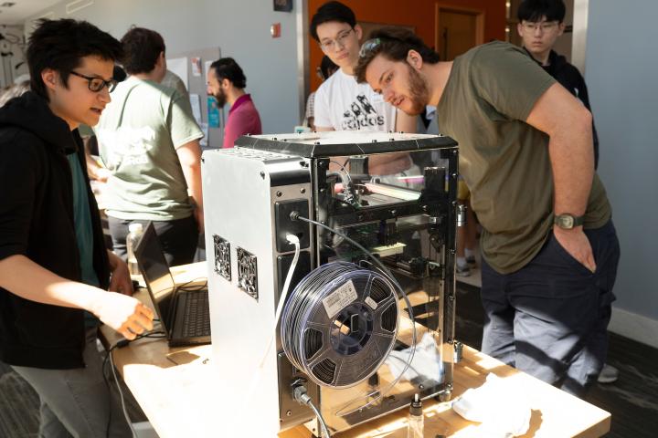 Students gather around a 3D printer during a project showcase.