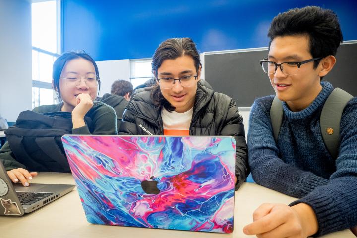 Three college students smile while sitting in a classroom and looking at a laptop screen at Olin College of Engineering, in Needham, MA.