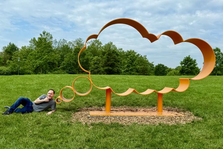 Jonathan Adler poses next to a thought bubble sculpture in a field. 