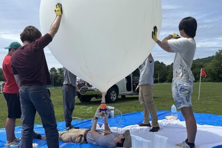 Olin students are holding up a large, white, high-altitude weather balloon as another team member lays flat on the ground inflating it. 