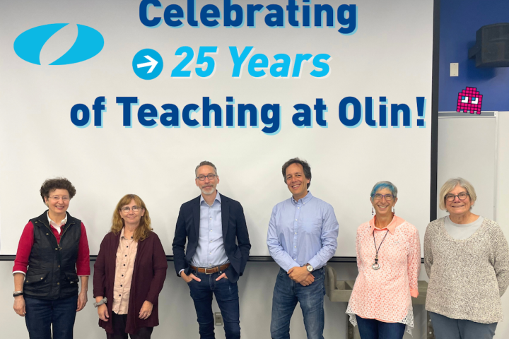 A group of six Olin College faculty members pose for a photo in a classroom with the embedded text behind them reading: "Celebrating 25 Years of Teaching at Olin!"