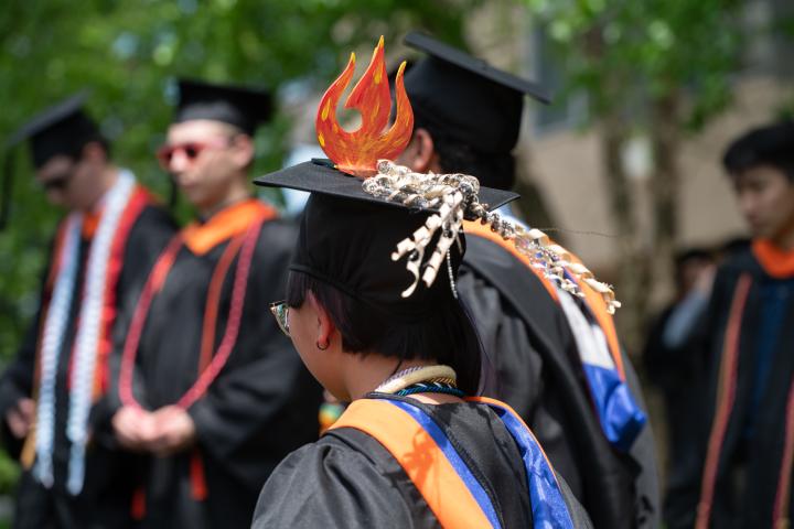 Commencement 2025, students in cap and gowns. Students fire decorated hat.