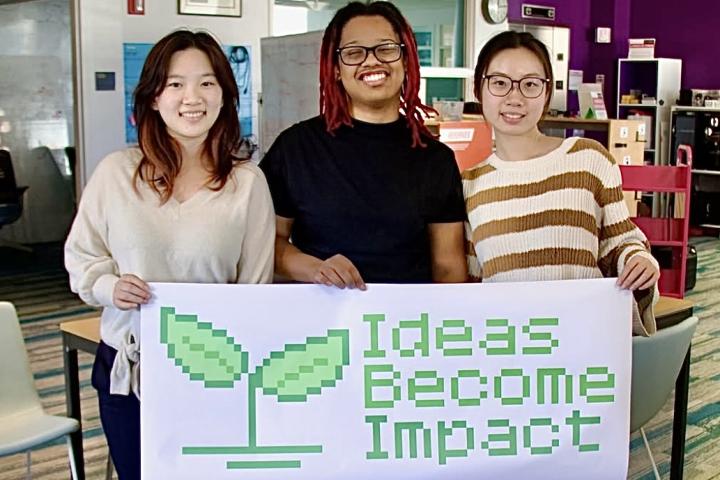 Three students stand with a white banner that has a green sprout and "Ideas Become Impact." 