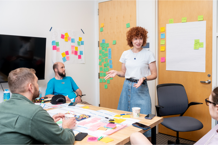 Three people sitting around a table. A fourth person stands behind the table gesturing, sticky notes adorn the table and nearby walls.
