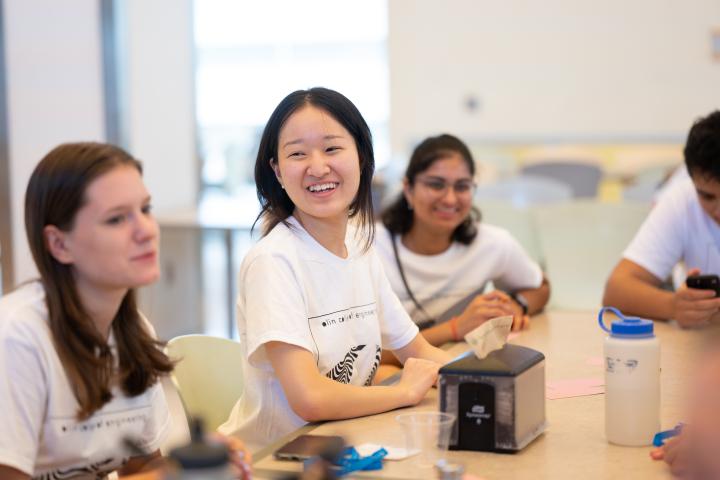 An Olin College of Engineering student is wearing a white "Olin" shirt and smiling while sitting with friends in the Campus Center cafe.