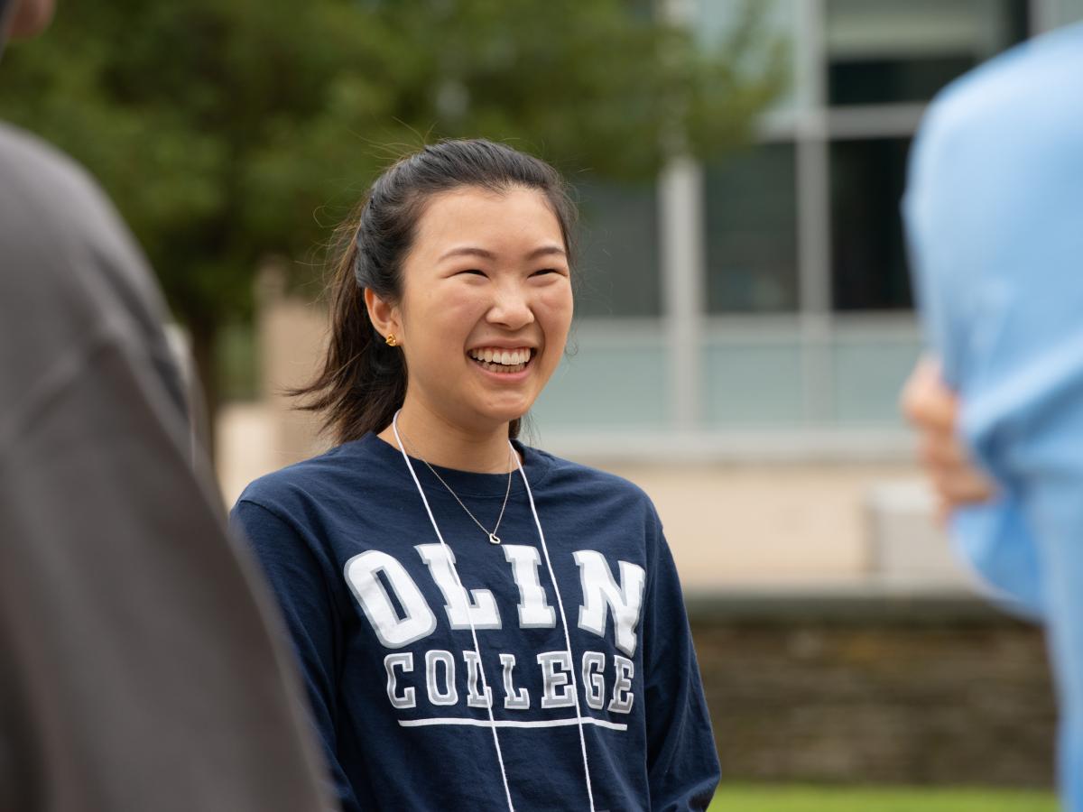 A dark haired student with a navy "Olin College" sweatshirt smiles candidly for the camera while on Olin's campus.