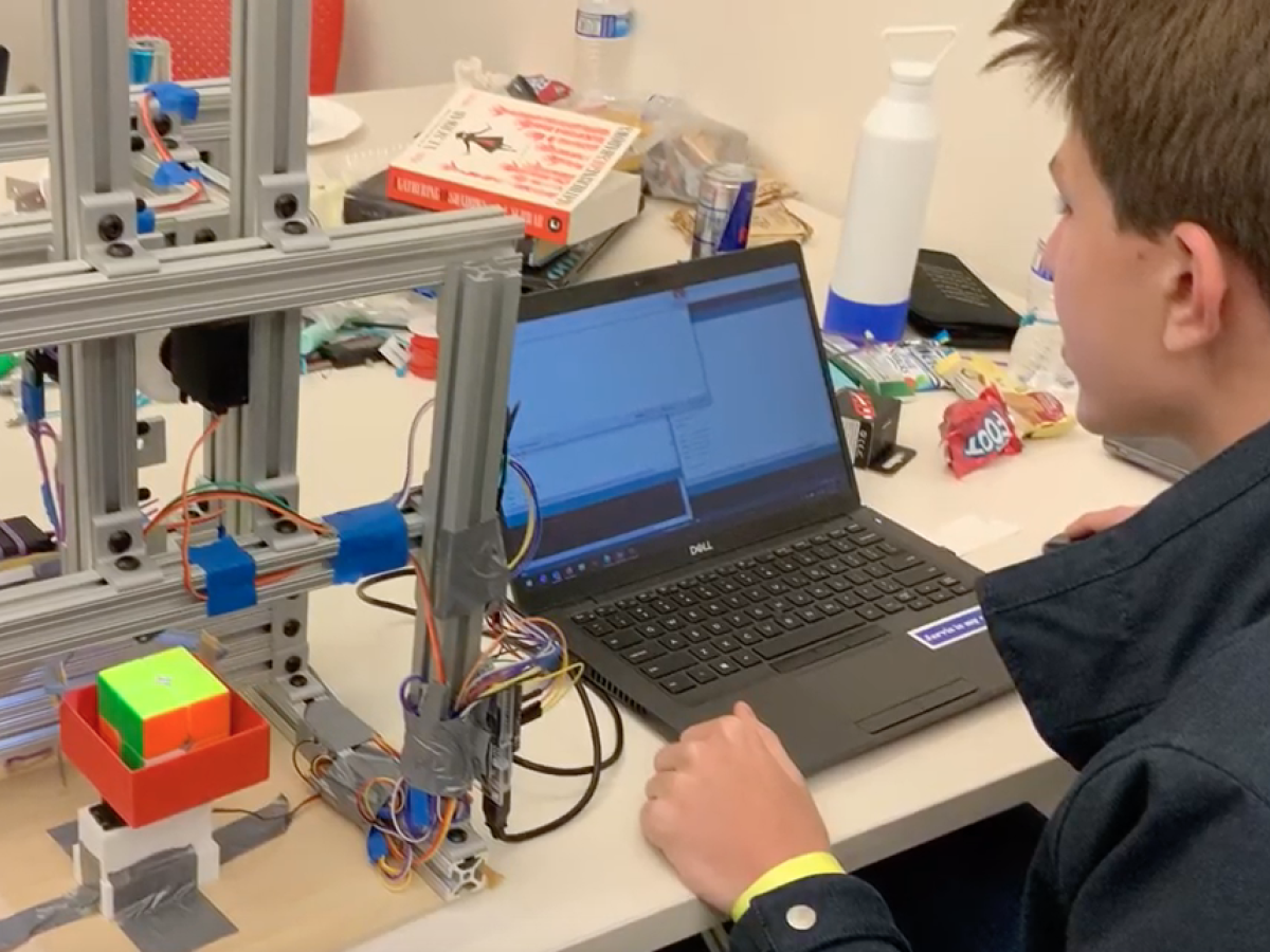 Photo/video still of the 2x2x2 Rubix Cube Solver. Photo by Nabih Estefan Diaz ’23. A student with black puffy vest, with back to the camera, working on a laptop at a table.
