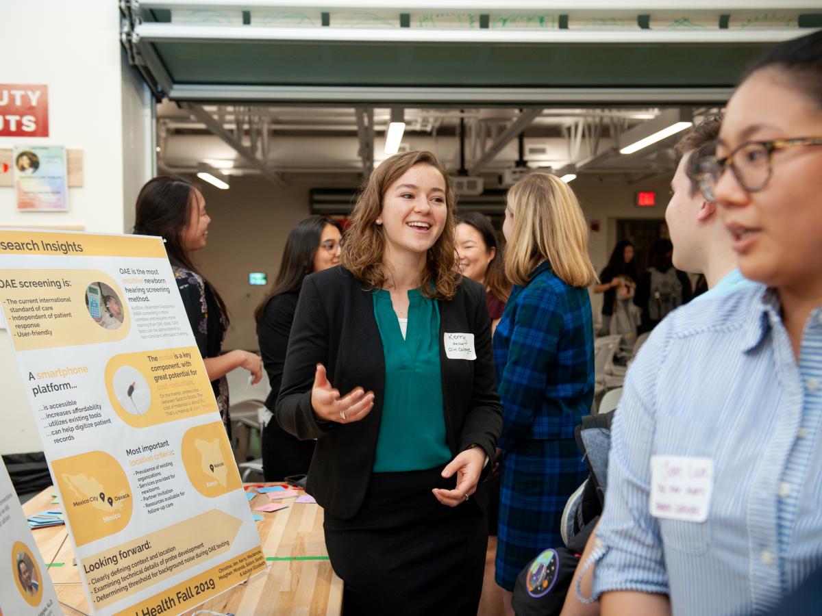 An ADE in green blouse and black blazer student talks with guests during poster presentation.