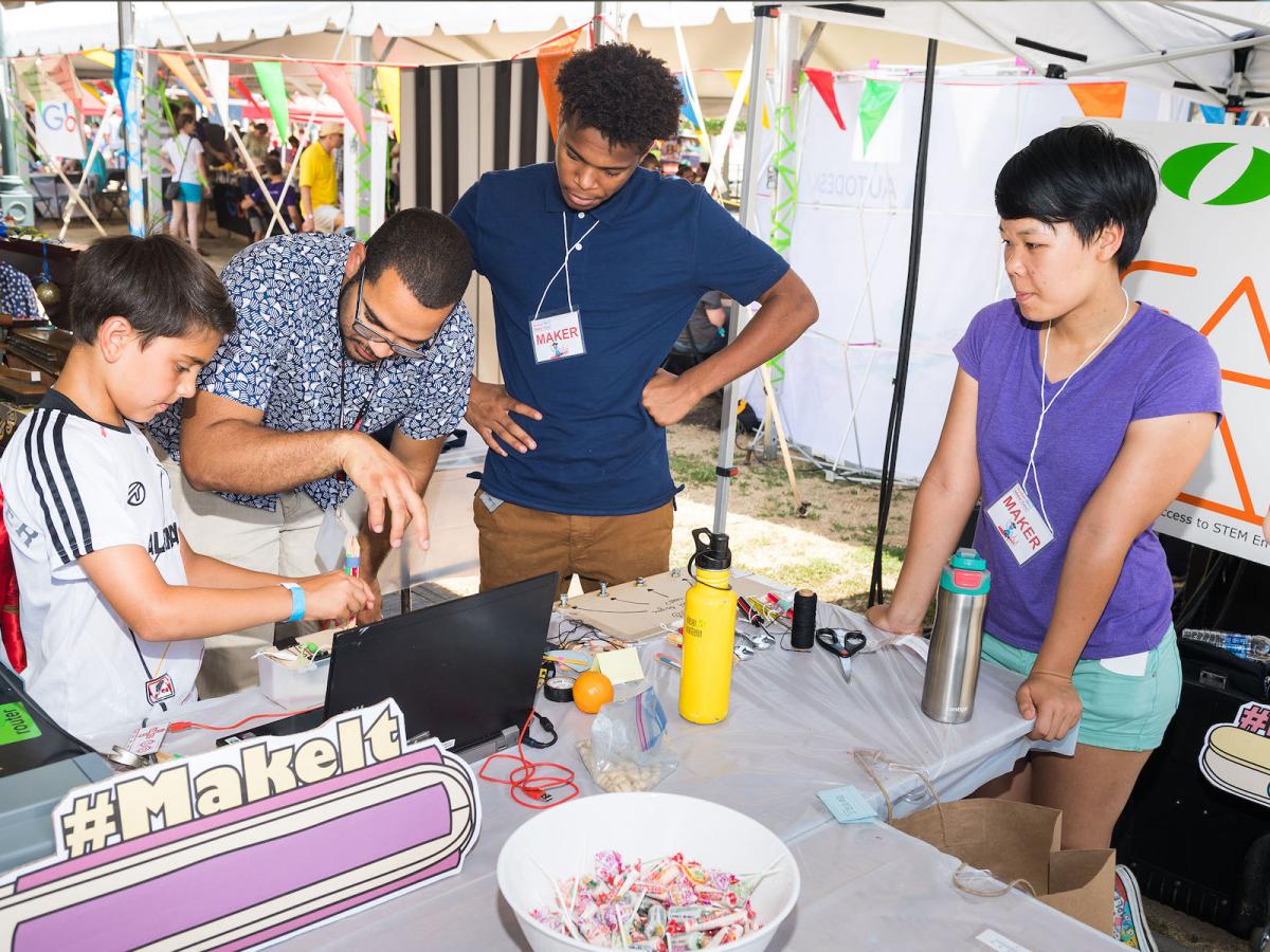 Professor Amon Millner helps a community member with a DIY STEM project as part of the STEAMing it in the Park series funded in part by the Boston Foundation. A man helps a young boy with a DIY coding project with a laptop and a tool made out of popsicle sticks with people looking on.
