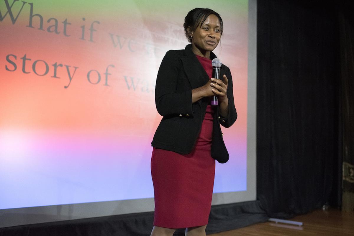Etosha Cave in black blazer and red dress holds a mic while presenting on stage.