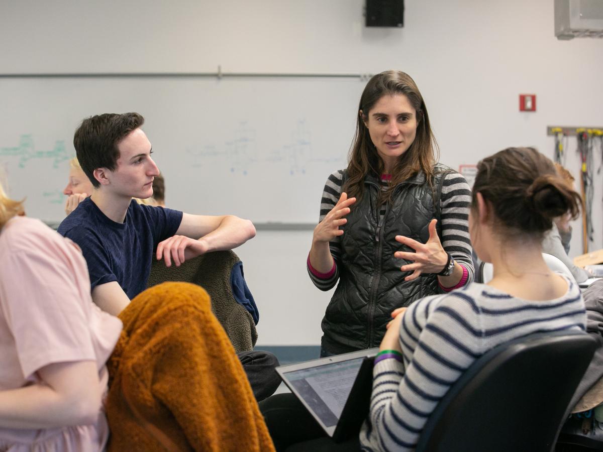 A professor in black vest and striped shirt, speaks to two students in a classroom.