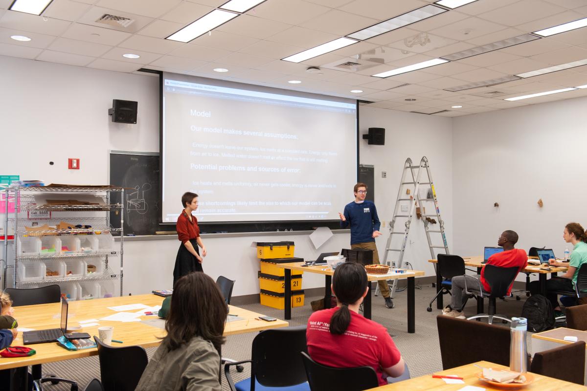 Students in a recent ModSim class present at the front of a classroom with screen down and ladder extended next to them.