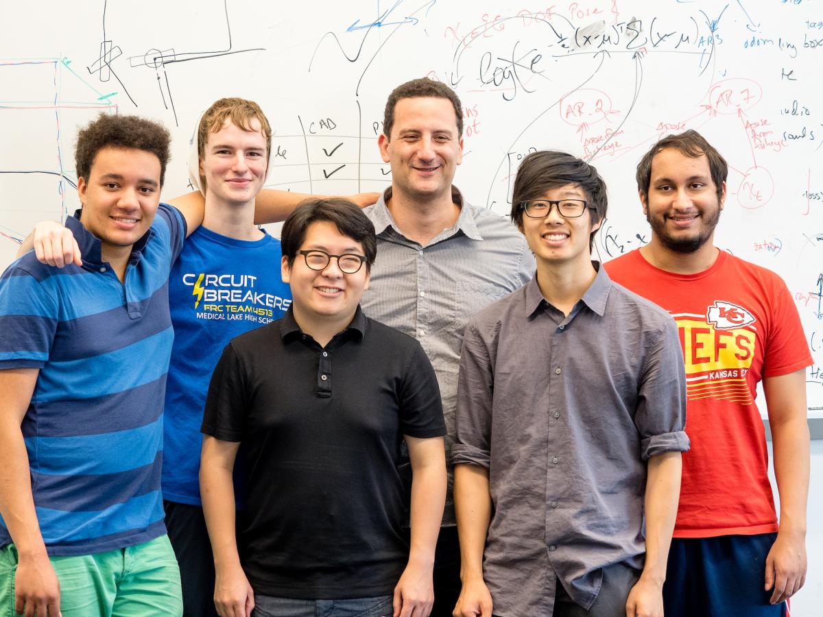 Paul Ruvolo, Associate Professor of Computer Science, poses with a group of five students in front of a whiteboard.