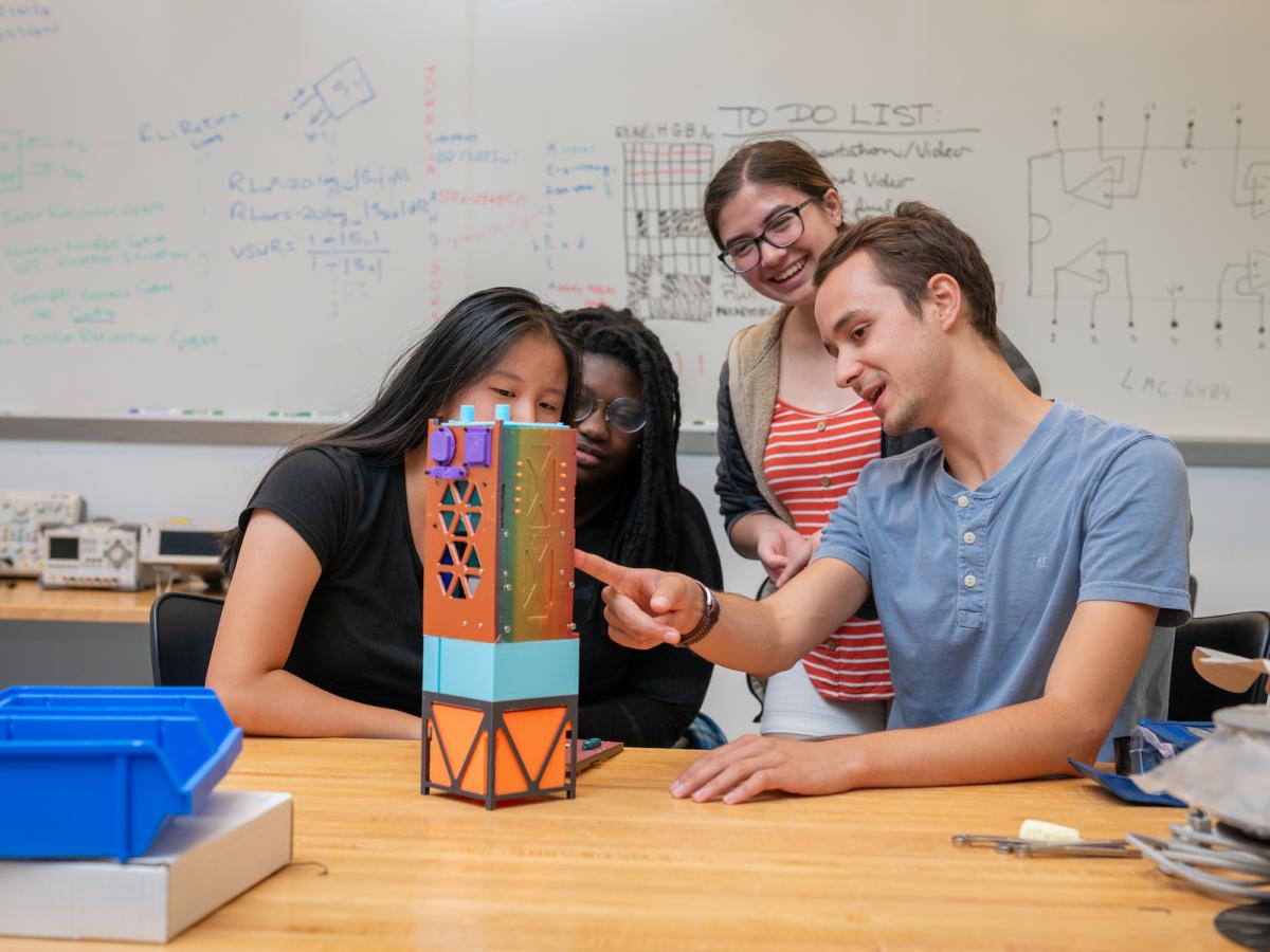Students from the Olin Satellite + Spectrum Technology & Policy (OSSTP) Group work this past summer on a prototype of one of the CubeSats. Four students work together in a classroom, pointing to a multicolored Cube prototype.