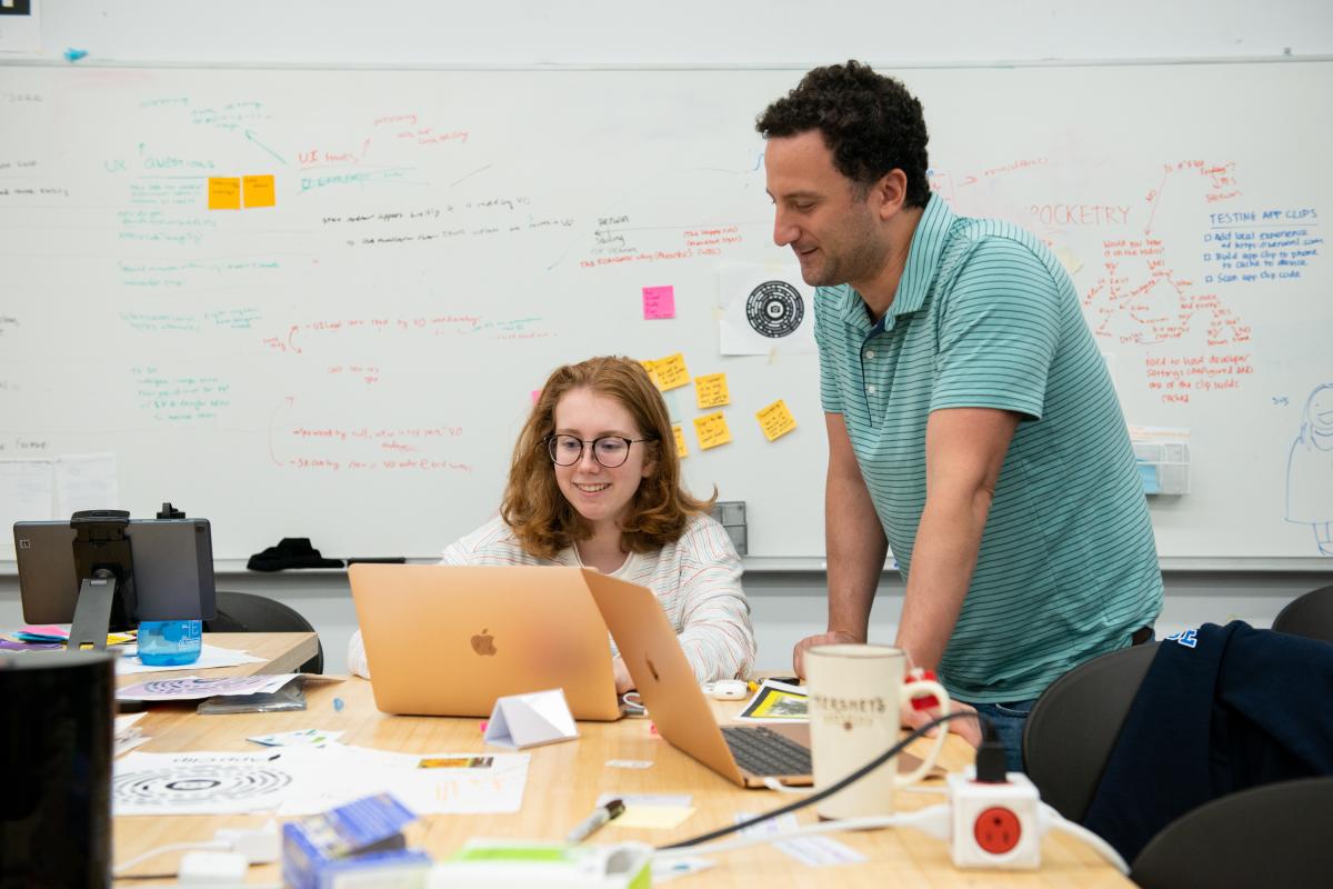 A photo of a professor and a student in a classroom looking at a laptop computer