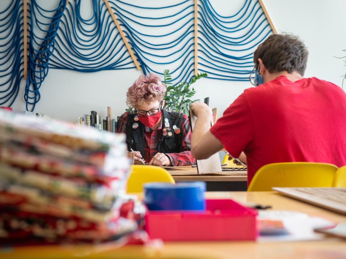 Two students work at a table, with multi-colored cloth in the foreground and a decorative rope wall hanging in background.