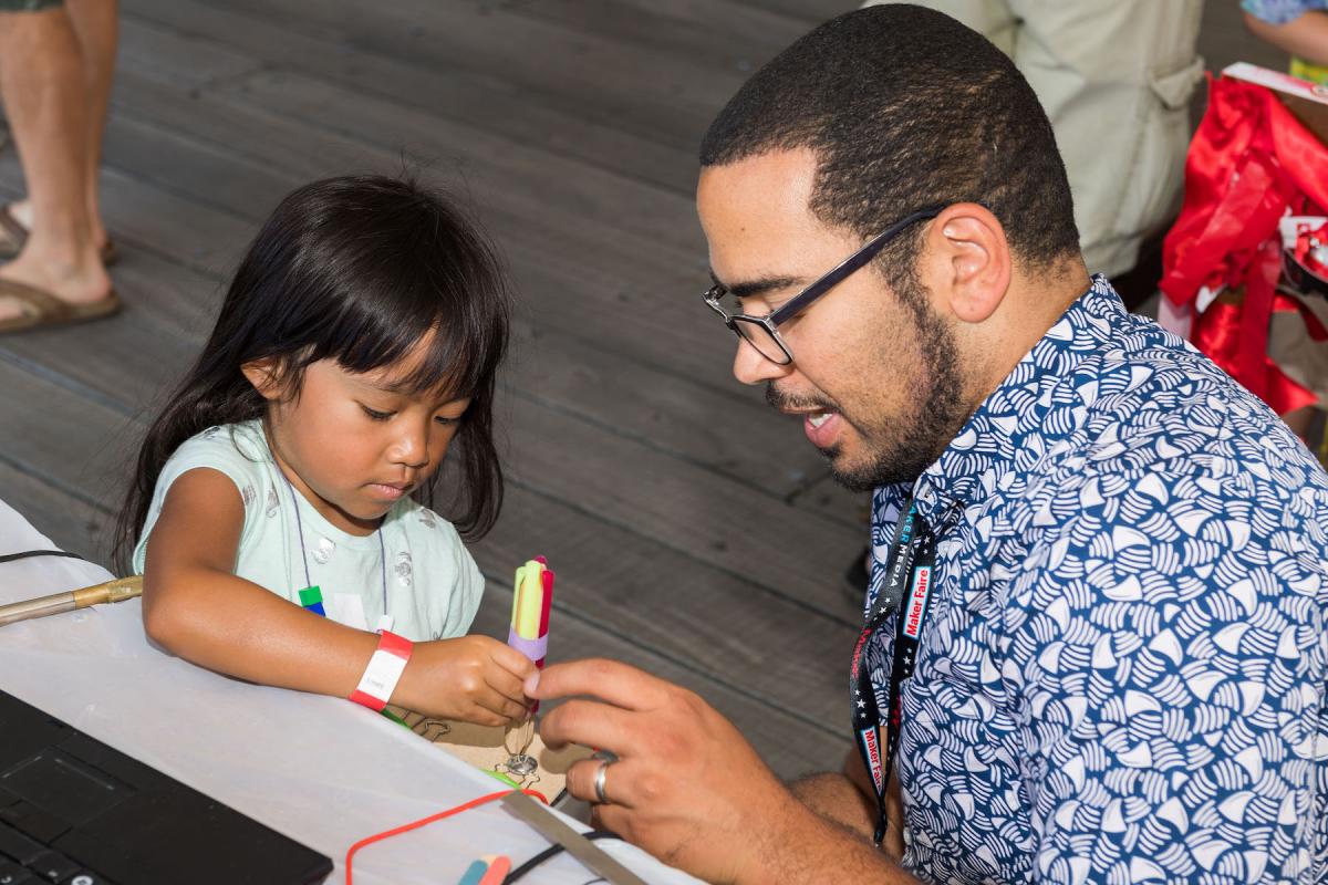 A man with a blue and white shirt and glasses helps a young girl draw on paper.