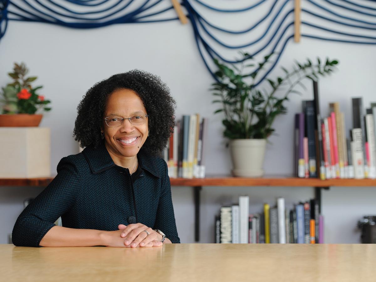 Olin College President Gilda A. Barabino, in a dark top and glasses, sits at a wooden table and smiles for the camera.