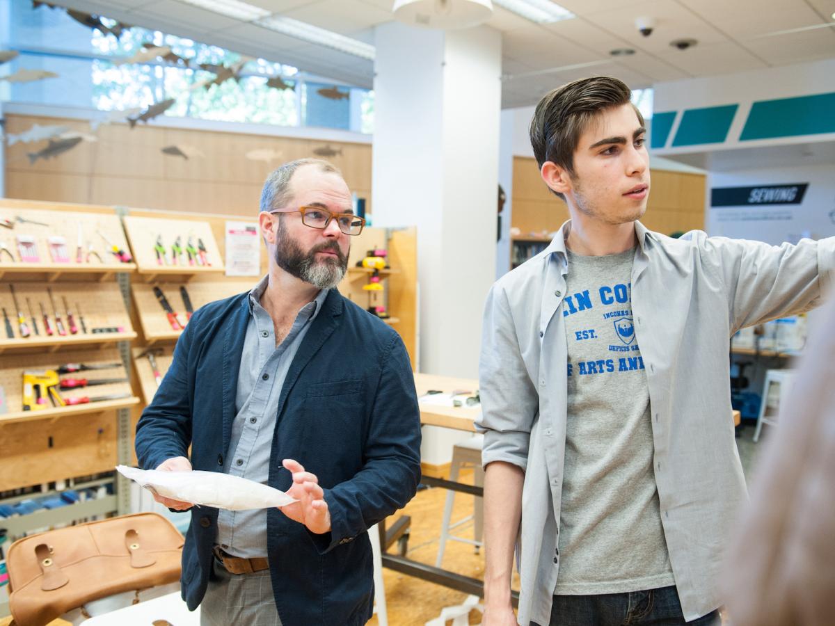 Bennett Taylor '22 looks over the show costumes he designed as part of a special collaboration with Tom Pearson, Olin's Sketch Model Creative-In-Reference for academic year 2019 - 2020. Two people in a library look at something on a wall off camera.