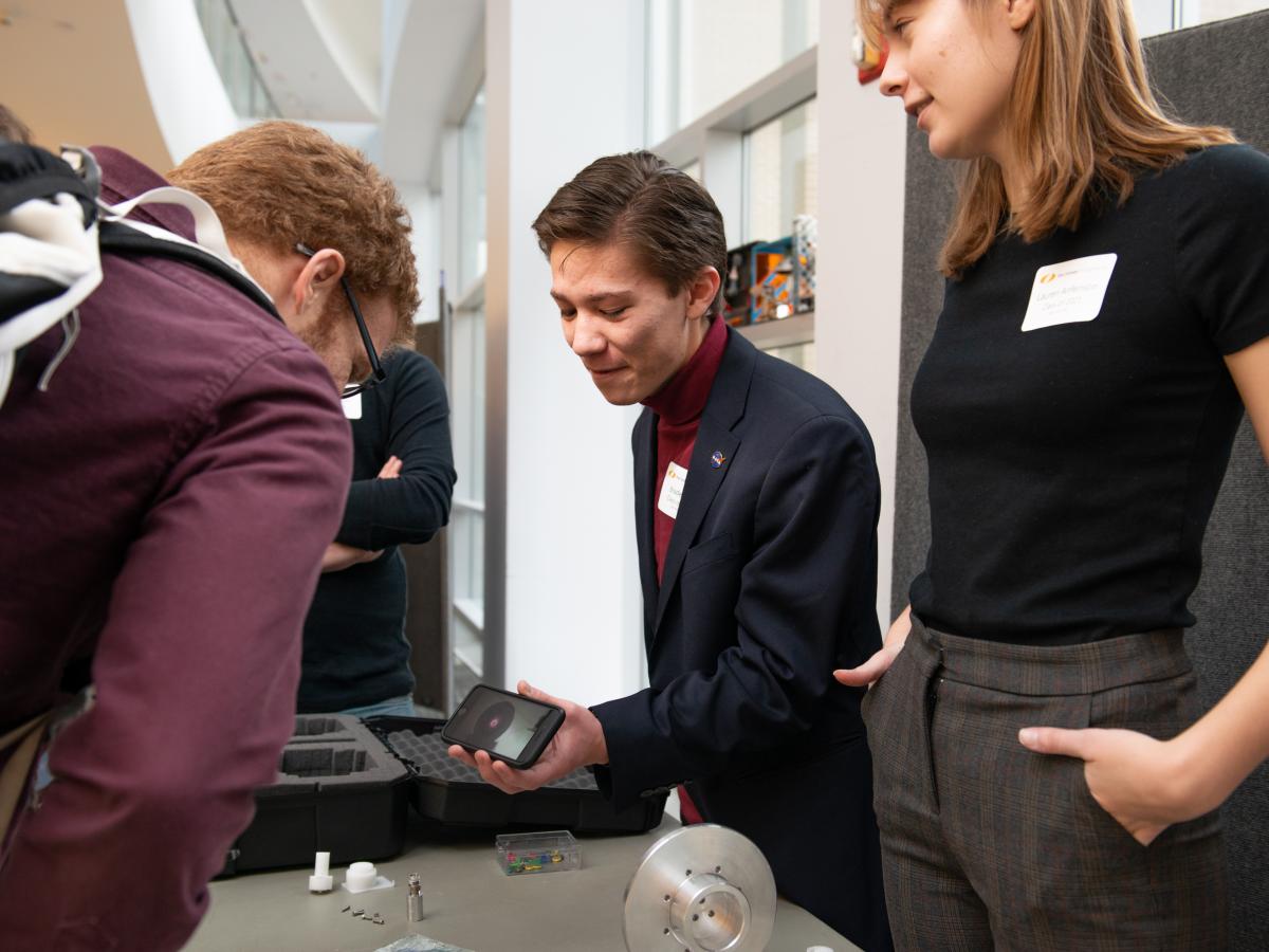 Braden Oh (L) and Lauren Anfenson (R) show a smartphone screen to an attendee in the Academic Center during Expo. Two students - both in black tops, stand behind a table and show another person in maroon shirt and backpack a smartphone screen.