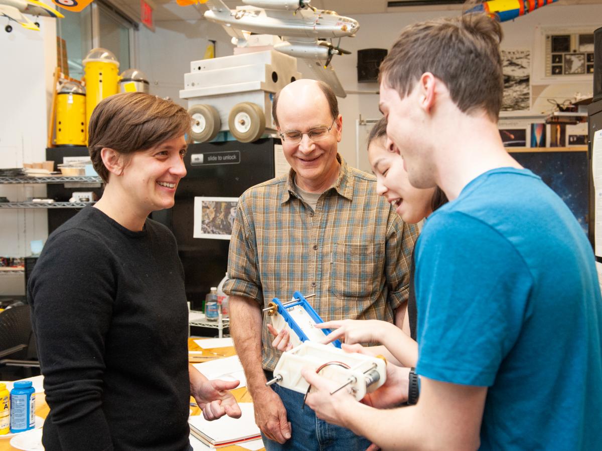 Professors Dave Barrett (Center) and Daniella Faas (Left) talk with students in 2019 Professor Dave Barrett (in the center) with checkered shirt and glasses, and Professor Daniella Faas (on the left) in black shirt and close-cropped hair, talk with students in a machine lab in 2019.