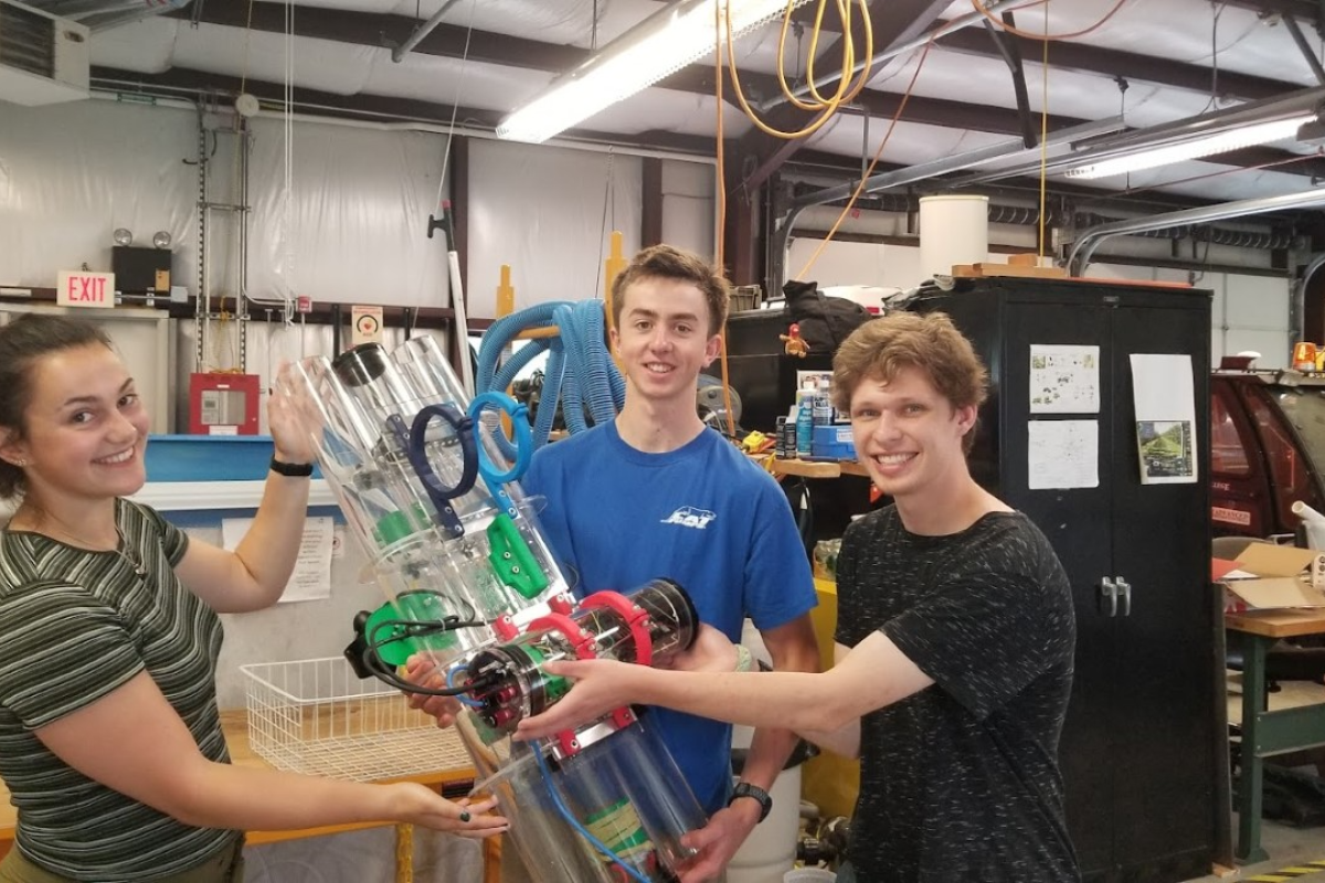 OCAP team members Zoie, Shawn, and Sander hold up the team's robot in Olin’s Large Project Building (LPB). Three students hold up a large cylinder device with red and green parts sticking onto it's body.