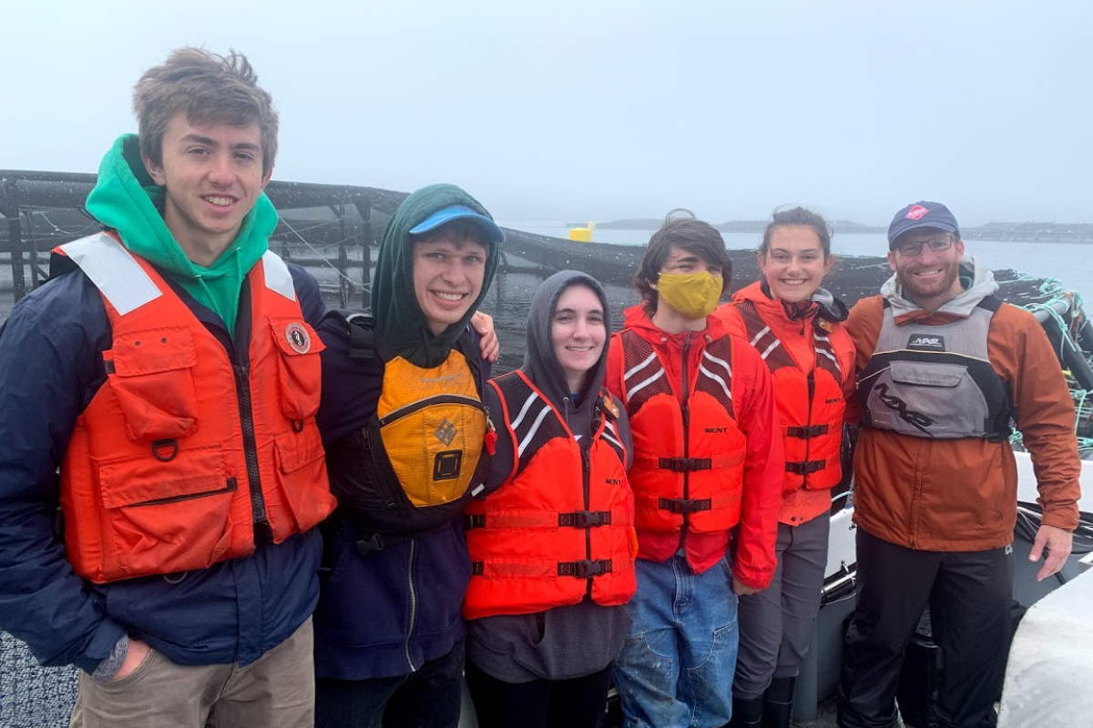 The OCAP team in Machiasport ME. (L to R): Shawn Albertson '21; Sader Miller '22; Sofia Goldberg '24; Lilly Novak '22; Zoie Leo '24; Professor Jeff Dusek. A team of five students and one professor line up, all wearing orange life preserver vests with the ocean in the background.