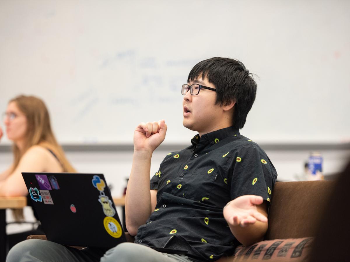 A man with glasses and dark hair, seated in a classroom with a laptop resting on his lap, talks to the class. 