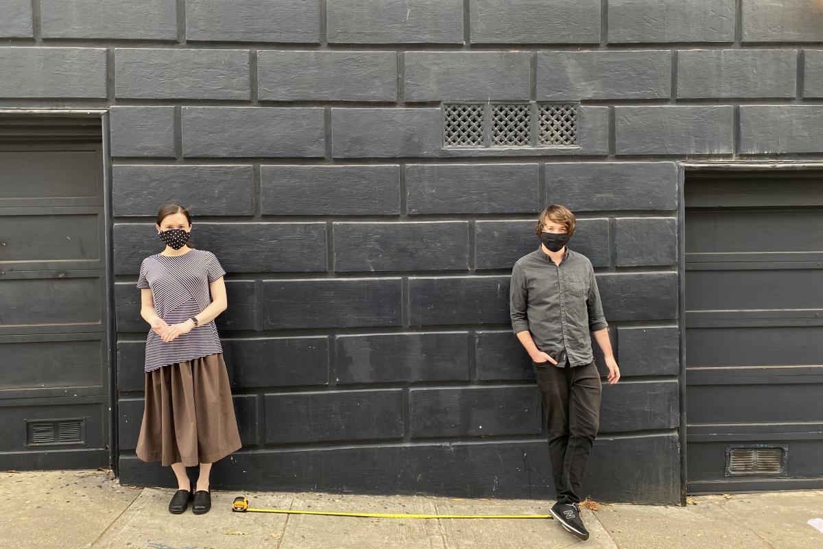 Two people stand against a grey, brick wall, approx. 6 feet apart.
