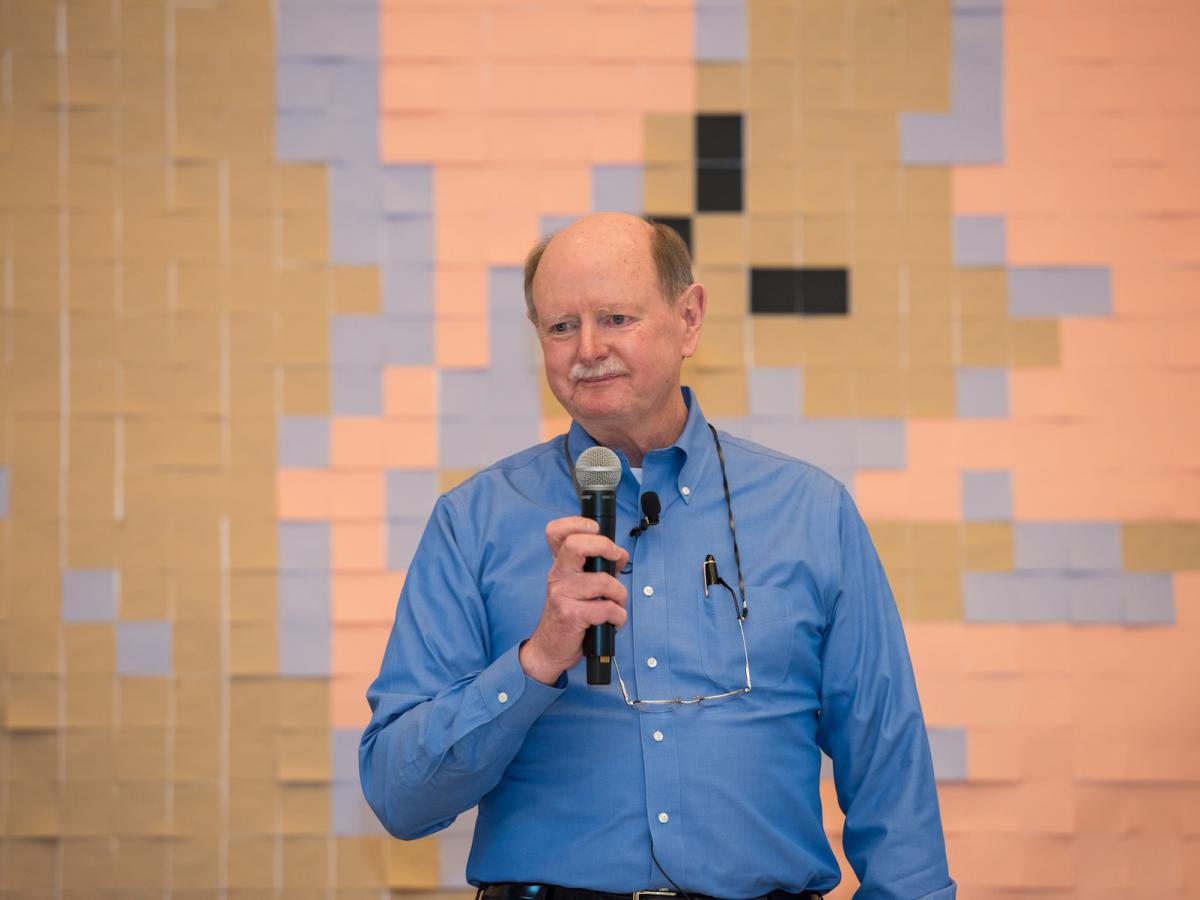 Rick Miller, Olin's first president, speaks during his retirement celebration. Olin's first president Rick Miller in blue dress shirt holds a mic in front of a wall of peach sticky notes.
