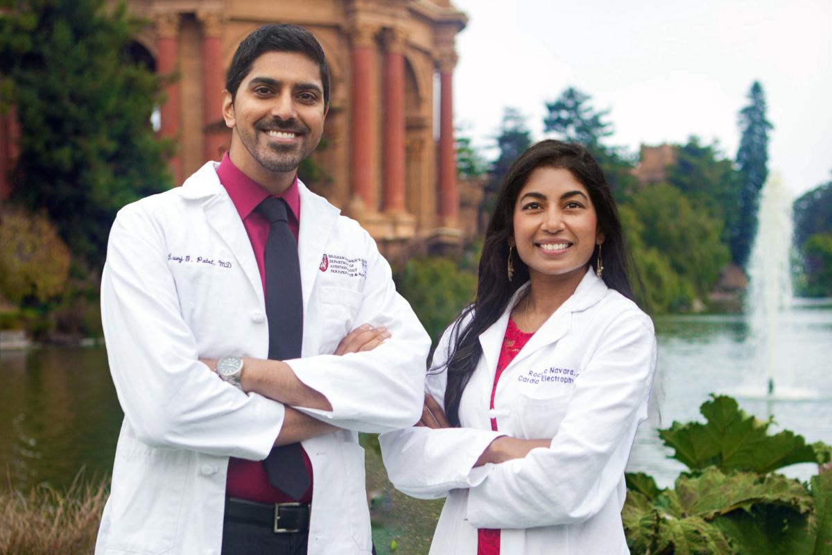 Dr. Rachita Navara ’11 poses with her husband and co-founder of SafeBeat Rx, Dr. Kunj Patel. A man in white lab coat and tie showing underneath stands with arms crossed, smiling, next to a woman in white lab coat with crossed arms and smiling at camera..