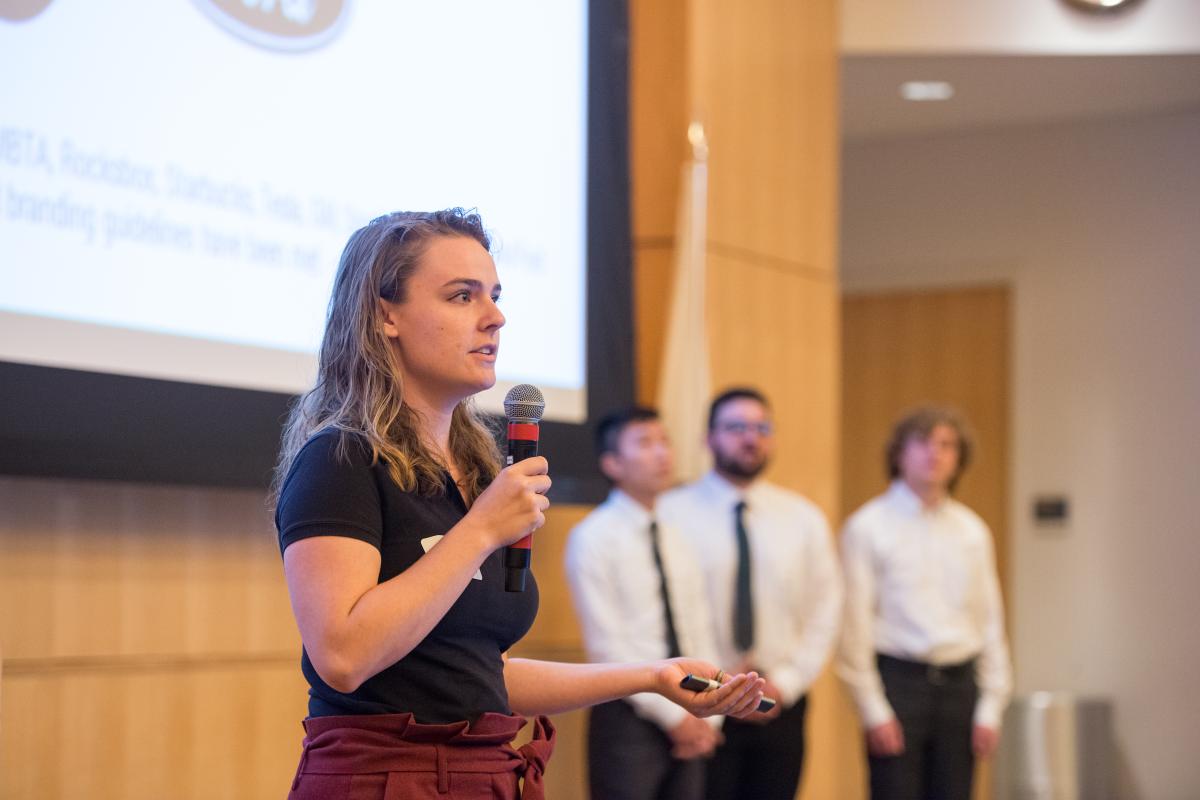 An Olin College student in black blouse and maroon pants holds a mic and presents in front of an audience of peers and corporate sponsors on May 2019 during Olin's annual SCOPE Summit.