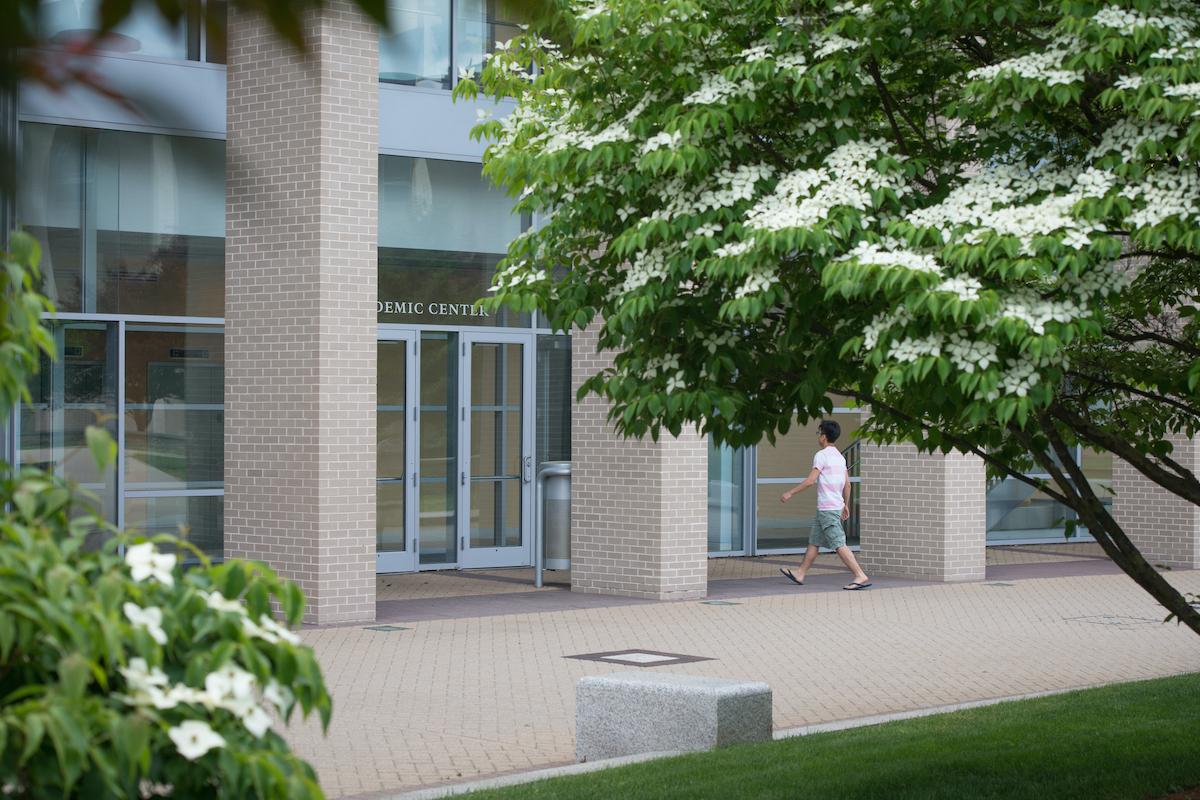 A student in pink shirt and shorts walks into the Academic Center building with green tree branches encroaching into the frame.