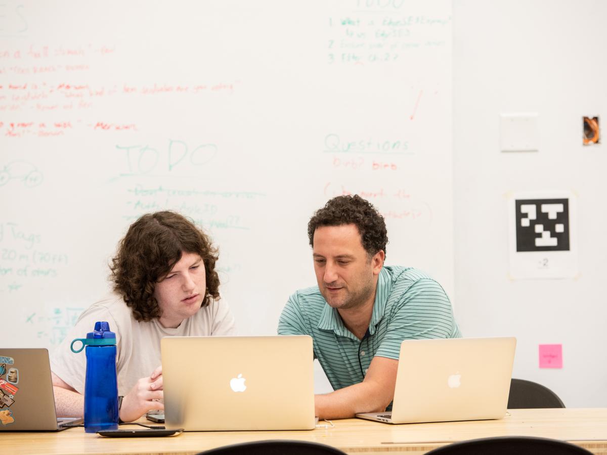 Associate Professor Paul Ruvolo in green shirt, works behind a laptop one-on-one with a student researcher, also behind a laptop, while sitting in a classroom.