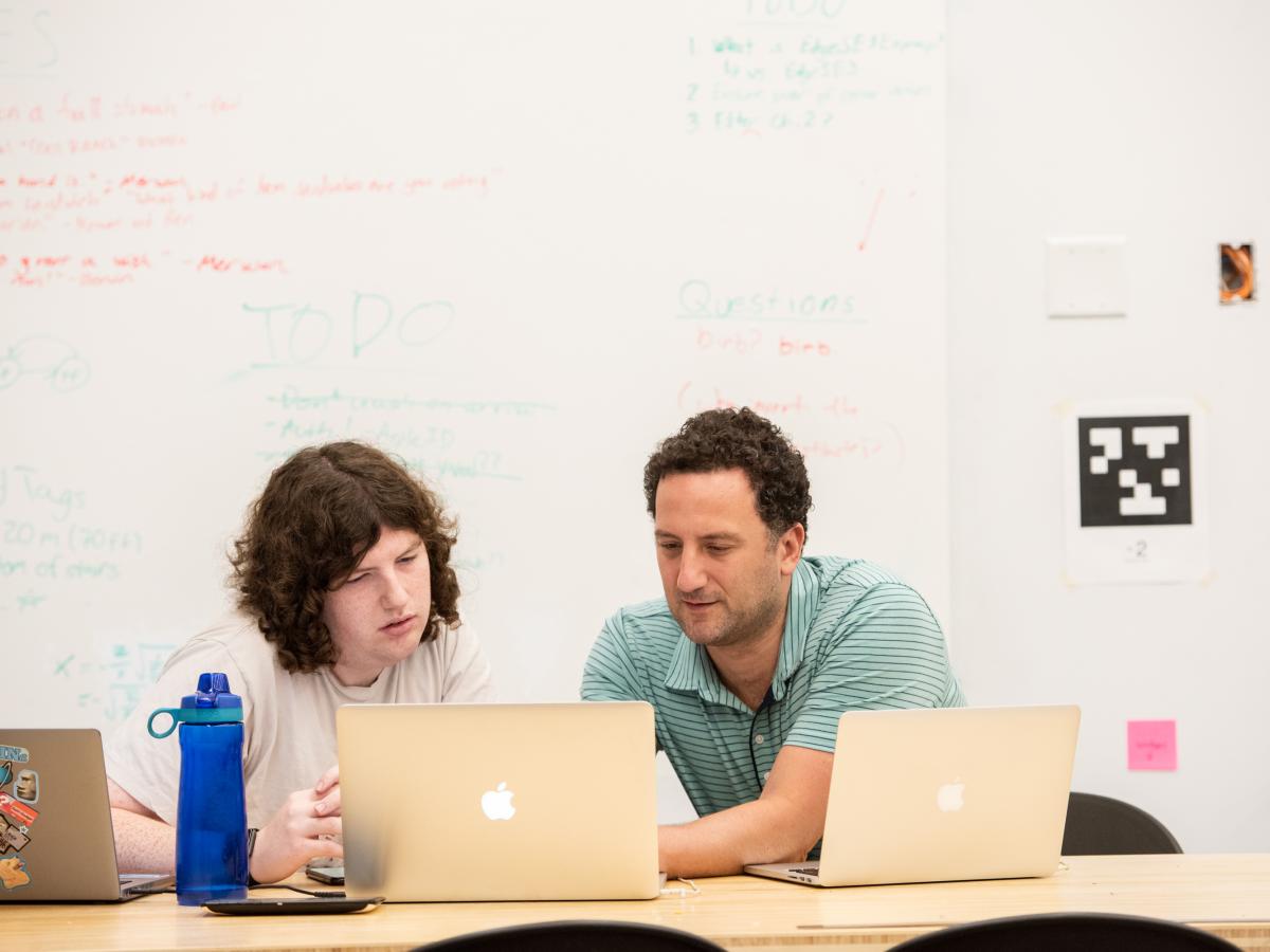Associate Professor Paul Ruvolo works one-on-one with a student researcher Associate Professor Paul Ruvolo in green shirt, works behind a laptop one-on-one with a student researcher, also behind a laptop, while sitting in a classroom.