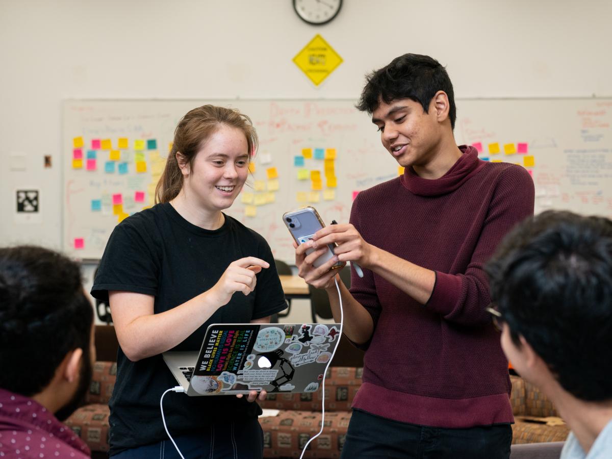 Summer researchers connect the phone and app to a laptop as the team looks on. Two summer research students in dark clothing connect a phone and app to a laptop via a white cord as two other team members look on.