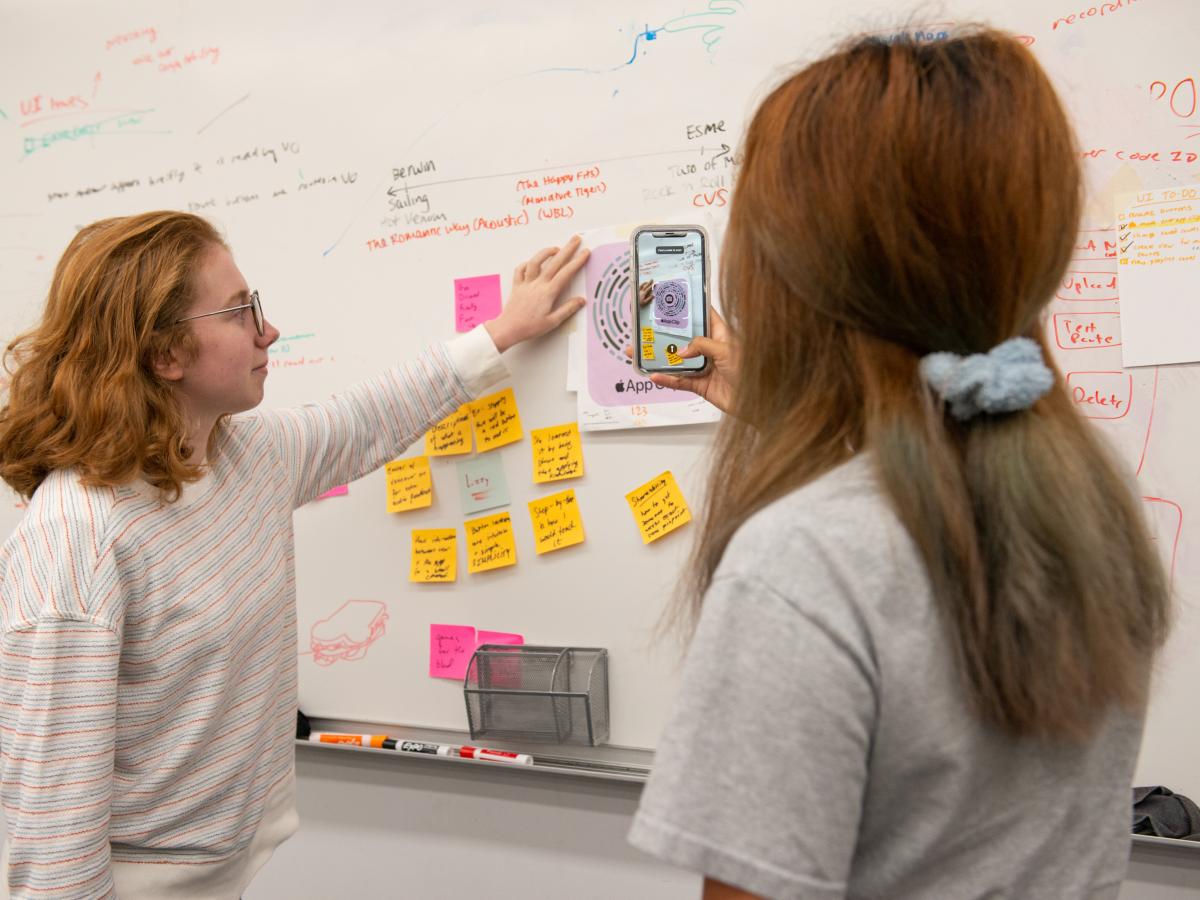 Olin students Esme '24 and Berwin '24 hold up and point a smartphone camera at a code on a whiteboard to use the assistive technology app Clew.