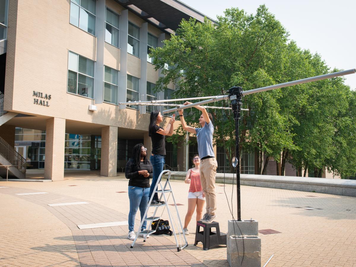 Four students with a ladder in the foreground work together to set up a large silver and black antenna. 