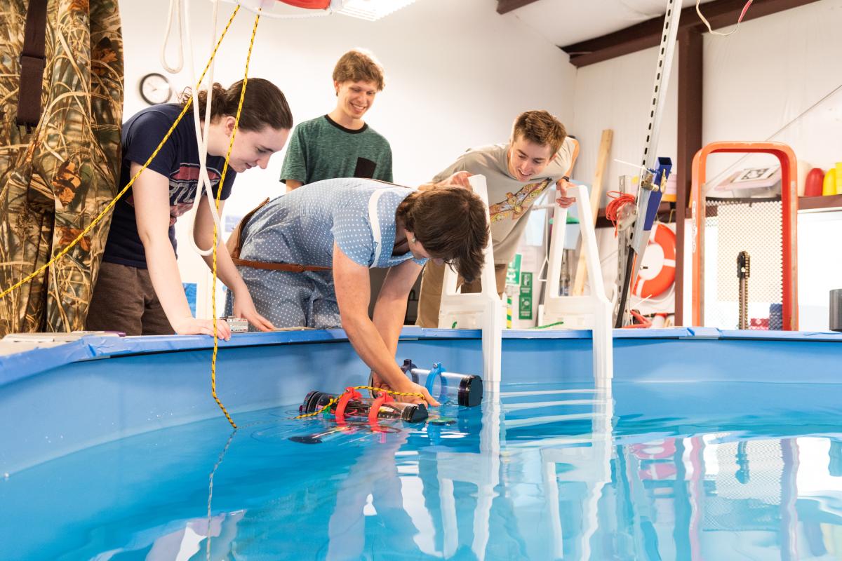 The research team tests the robot's up and down motions in Olin’s Large Project Building (LPB). Four student researchers hold down a small plastic device in a pool of water.