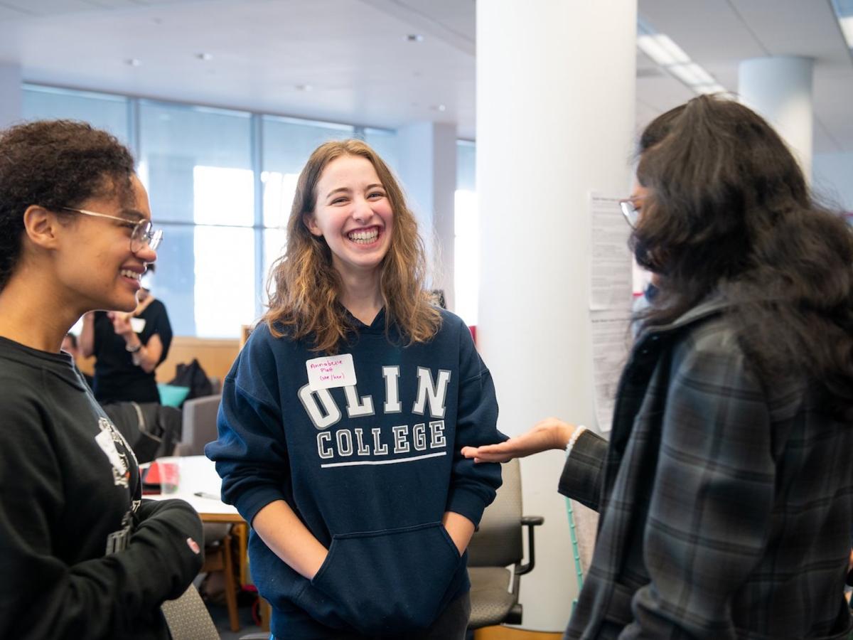 Students share a laugh together in the Olin Library during Build Week 2020. A student with a blue and white Olin College sweatshirt smiles while talking with two other people.