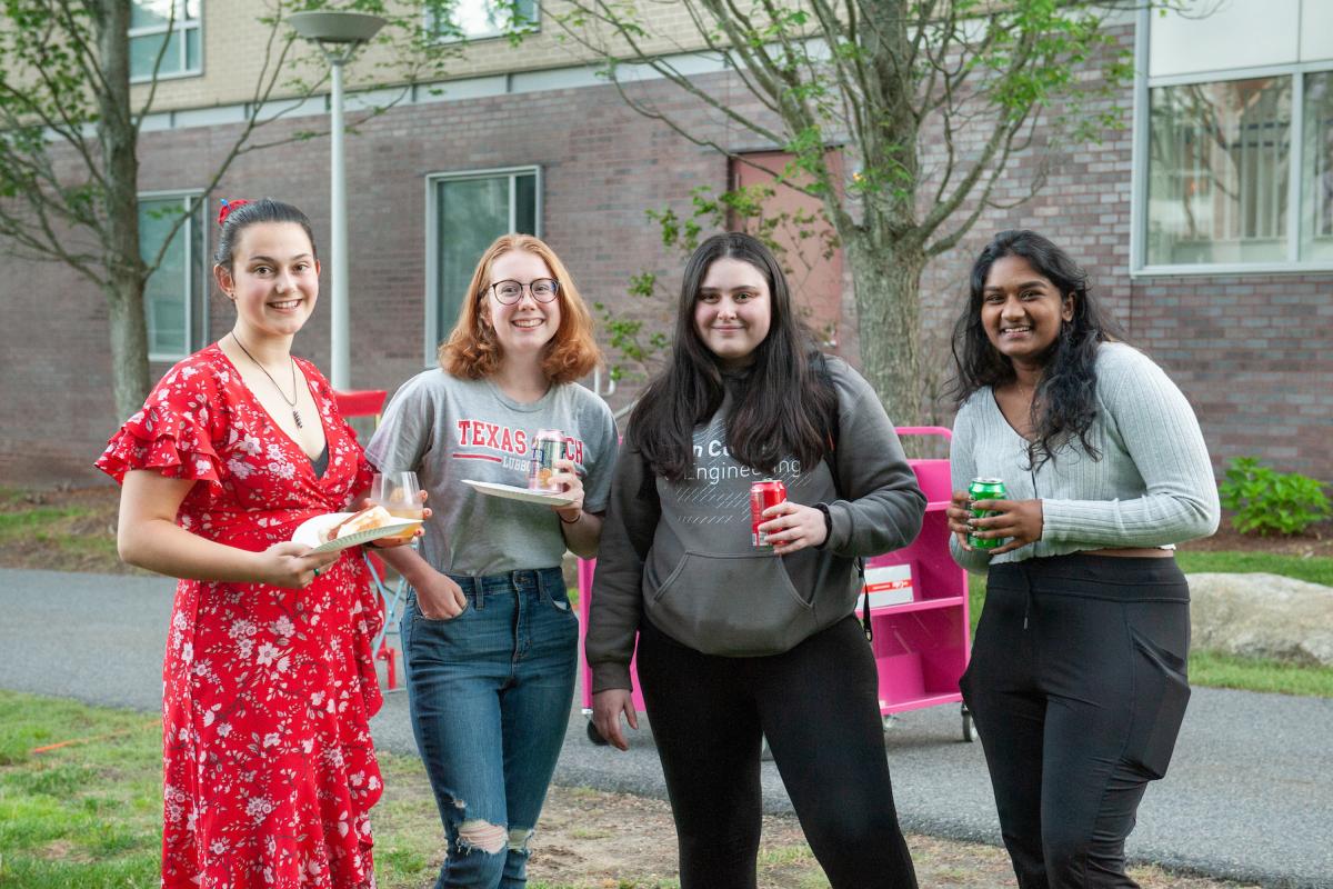 Four Olin students standing shoulder-to-shoulder, some with food and drink in hand, pose for a photo outside.