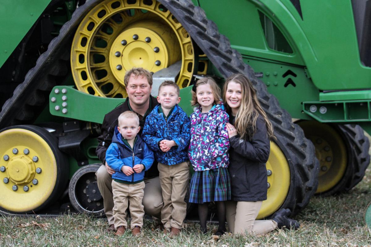 Clark McPheeters ’09 and Bryn (Hollen) McPheeters ’09 pose in front of a large green tractor with their three small children.