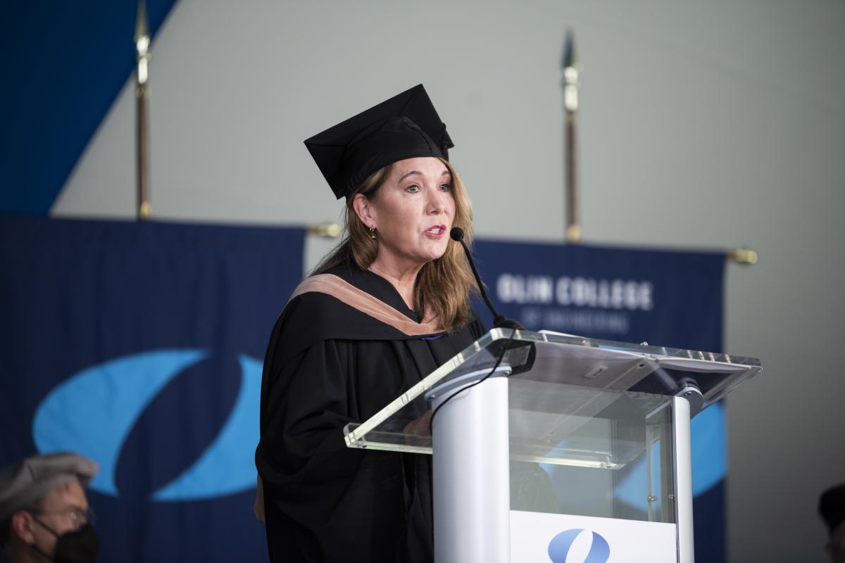 Beverly Wyse P’15, Chair of the Olin Board of Trustees, stands at a podium dressed in regalia, to welcome the graduates and their families during the Olin College 2022 Commencement Ceremony.