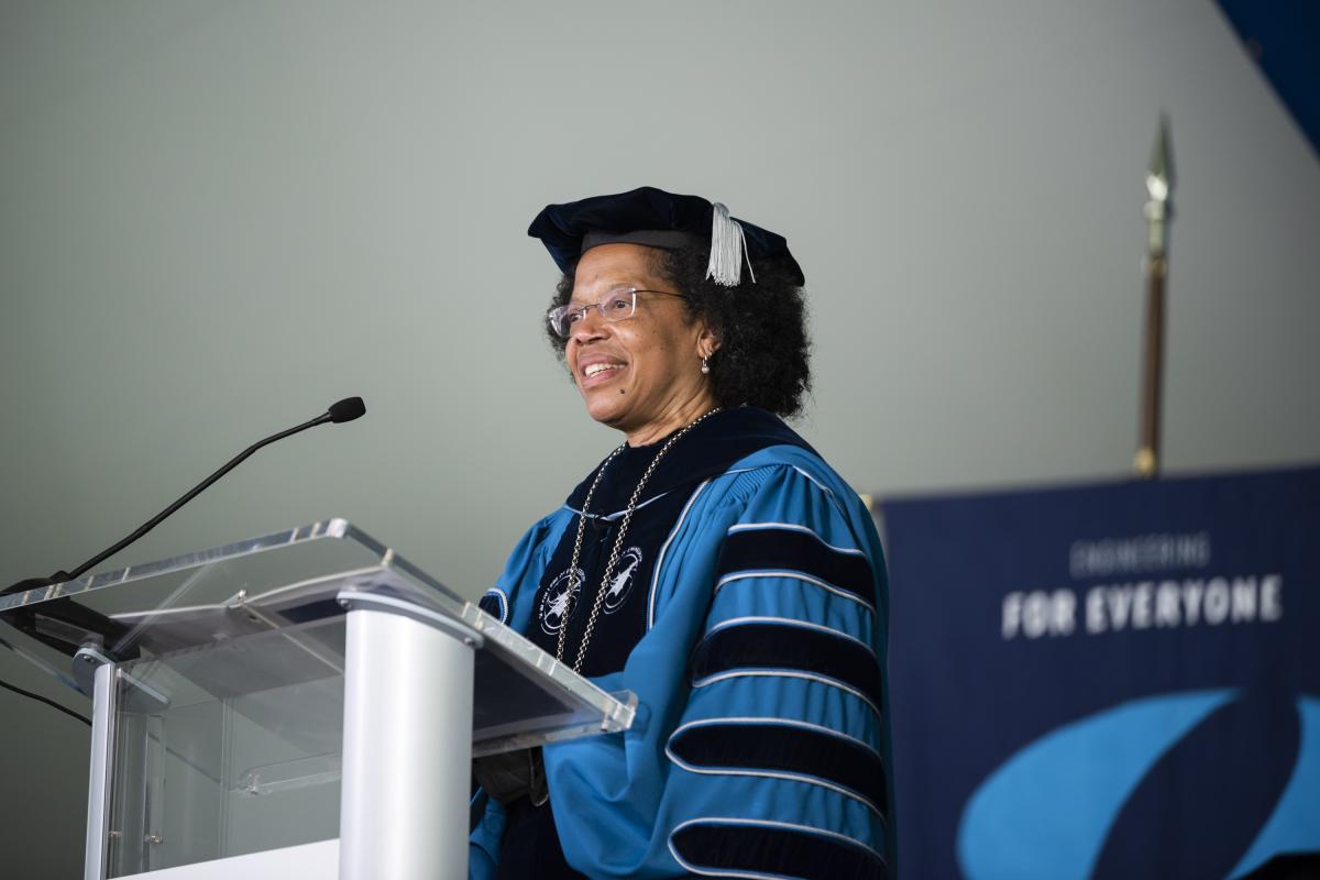 Olin College of Engineering President Dr. Gilda A. Barabino stands at the Commencement podium in graduation regalia to address the Class of 2022. Olin College of Engineering President Dr. Gilda A. Barabino stands at a podium dressed in blue and black graduation regalia.