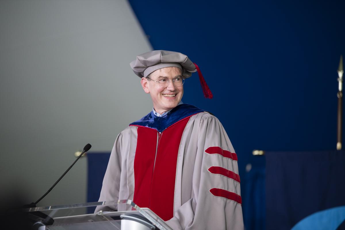 Provost Mark Somerville pictured at the podium wearing grey and red graduation regalia.