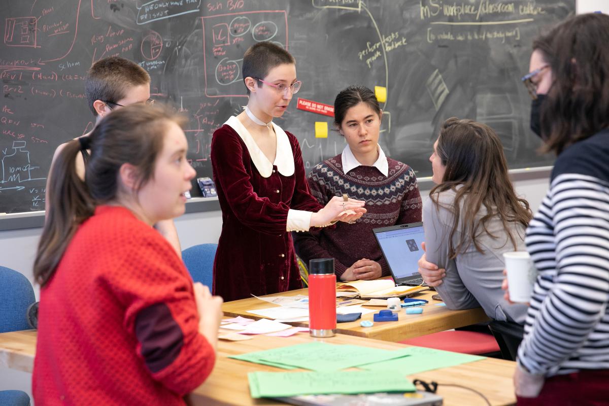 Multiple students collaborate with a professor around a table in a classroom with a large blackboard in the background.